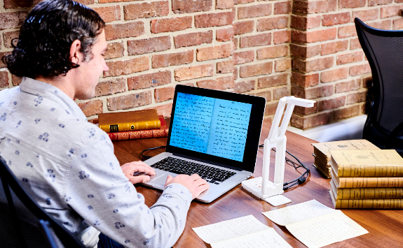 A man sat at a desk, in front of a laptop with a close up image of manuscript handwriting. Beside him on the desk is a visualiser looking over a manuscript.